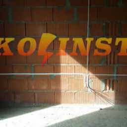 Białe przewody elektryczne prowadzone po ceglanej ścianie w surowym wnętrzu. Widoczny fragment podłogi w stanie deweloperskim. Na ścianie nałożone logo firmy.