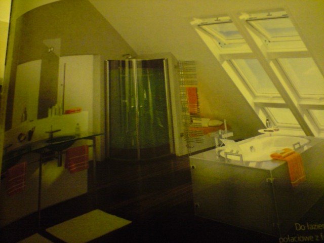 Jasna łazienka na poddaszu ze skośnym sufitem i oknami dachowymi, kabiną prysznicową, umywalką i wanną w obudowie z metalicznym wykończeniem. Pomarańczowe ręczniki dodają koloru.