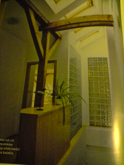 Wnętrze z drewnianą konstrukcją stropu, ścianą z luksferów i roślinami doniczkowymi na drewnianej zabudowie, doświetlone światłem z okien dachowych.