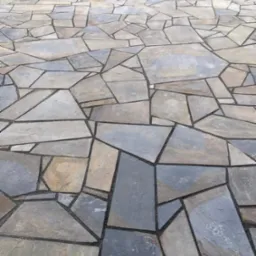 Nieregularny wzór ułożony z płaskich, wielokątnych płyt kamiennych w odcieniach szarości i beżu, tworzący powierzchnię tarasu lub ścieżki ogrodowej z widocznymi fugami wypełnionymi drobnym kruszywem.