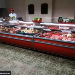 Długa lada chłodnicza w sklepie mięsnym, wypełniona różnymi rodzajami mięs i wędlin, z pracownikiem w tle i kafelkową podłogą.