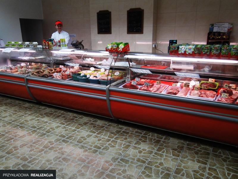 Długa lada chłodnicza w sklepie mięsnym, wypełniona różnymi rodzajami mięs i wędlin, z pracownikiem w tle i kafelkową podłogą.
