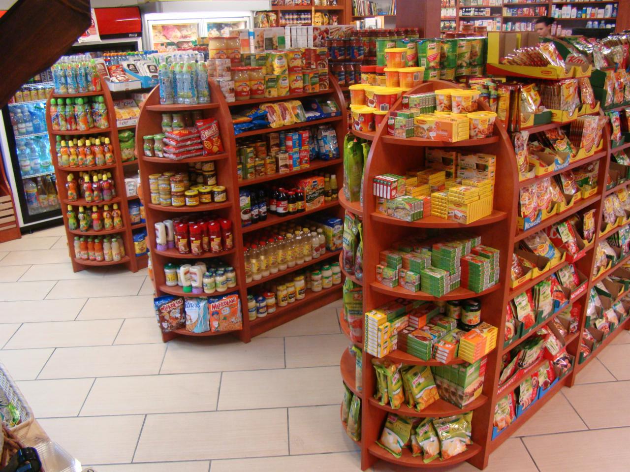 Wnętrze sklepu spożywczego z półokrągłymi regałami wypełnionymi różnorodnymi produktami: napojami, konserwami, przyprawami i słodyczami, widok na chłodziarkę z napojami w tle.