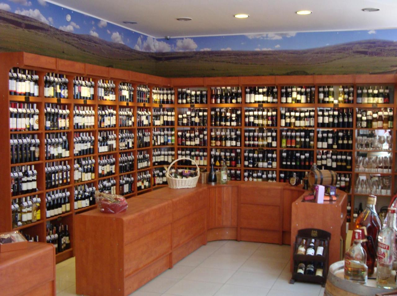 Wnętrze sklepu z winami: drewniane regały wypełnione butelkami wina, drewniana lada z koszem i dekoracjami, malowidło krajobrazu na ścianie nad regałami.