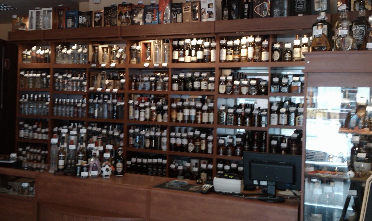 Drewniane regały sklepowe wypełnione różnorodnymi butelkami alkoholi, widoczne także ekspozytory za ladą i monitor komputera.