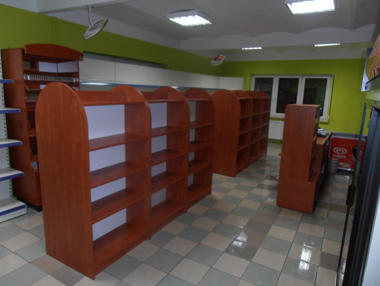 Wnętrze sklepu spożywczego z pustymi, drewnianymi regałami sklepowymi o zaokrąglonych krawędziach i białym tłem, widok w kierunku okna, szara podłoga z płytek.