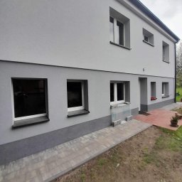 TOM-BUD USŁUGI OGÓLNOBUDOWLANE - Dwukondygnacyjny dom po remoncie elewacji, z nowym szarym tynkiem i ciemniejszym pasem u dołu, widoczny fragment chodnika z kostki brukowej i przygotowany teren pod ogród.