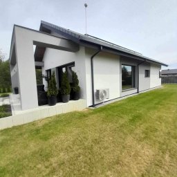 TOM-BUD USŁUGI OGÓLNOBUDOWLANE - Nowoczesny dom jednorodzinny z jasną elewacją, czarnymi ramami okiennymi i klimatyzatorem na ścianie, otoczony trawnikiem.