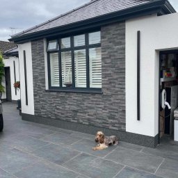 TOM-BUD USŁUGI OGÓLNOBUDOWLANE - Nowoczesny dom z elewacją z szarej cegły i białym tynkiem, czarnymi detalami oraz leżącym psem rasy Cocker Spaniel na szarym bruku.