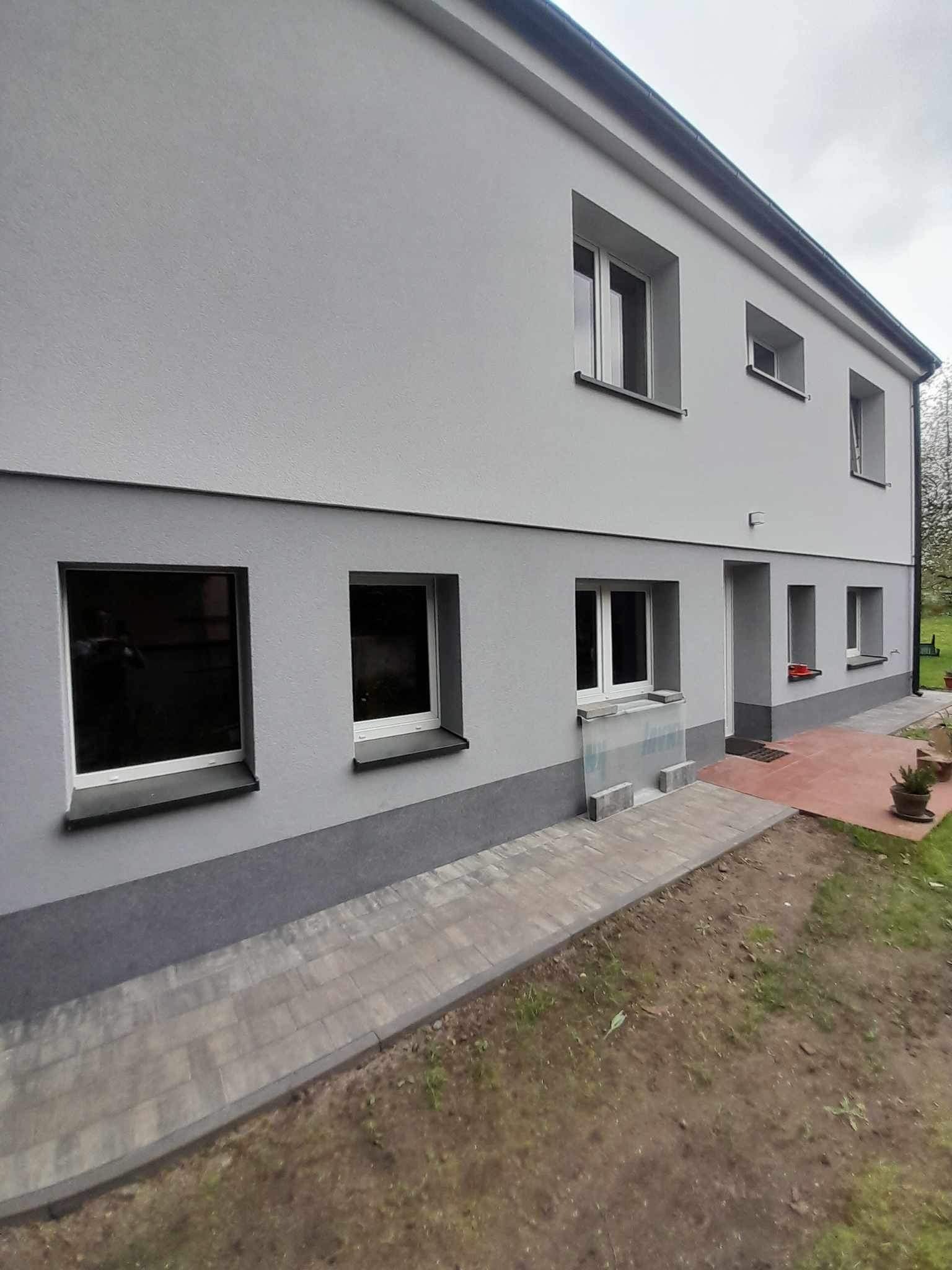 Dwukondygnacyjny dom po remoncie elewacji, z nowym szarym tynkiem i ciemniejszym pasem u dołu, widoczny fragment chodnika z kostki brukowej i przygotowany teren pod ogród.