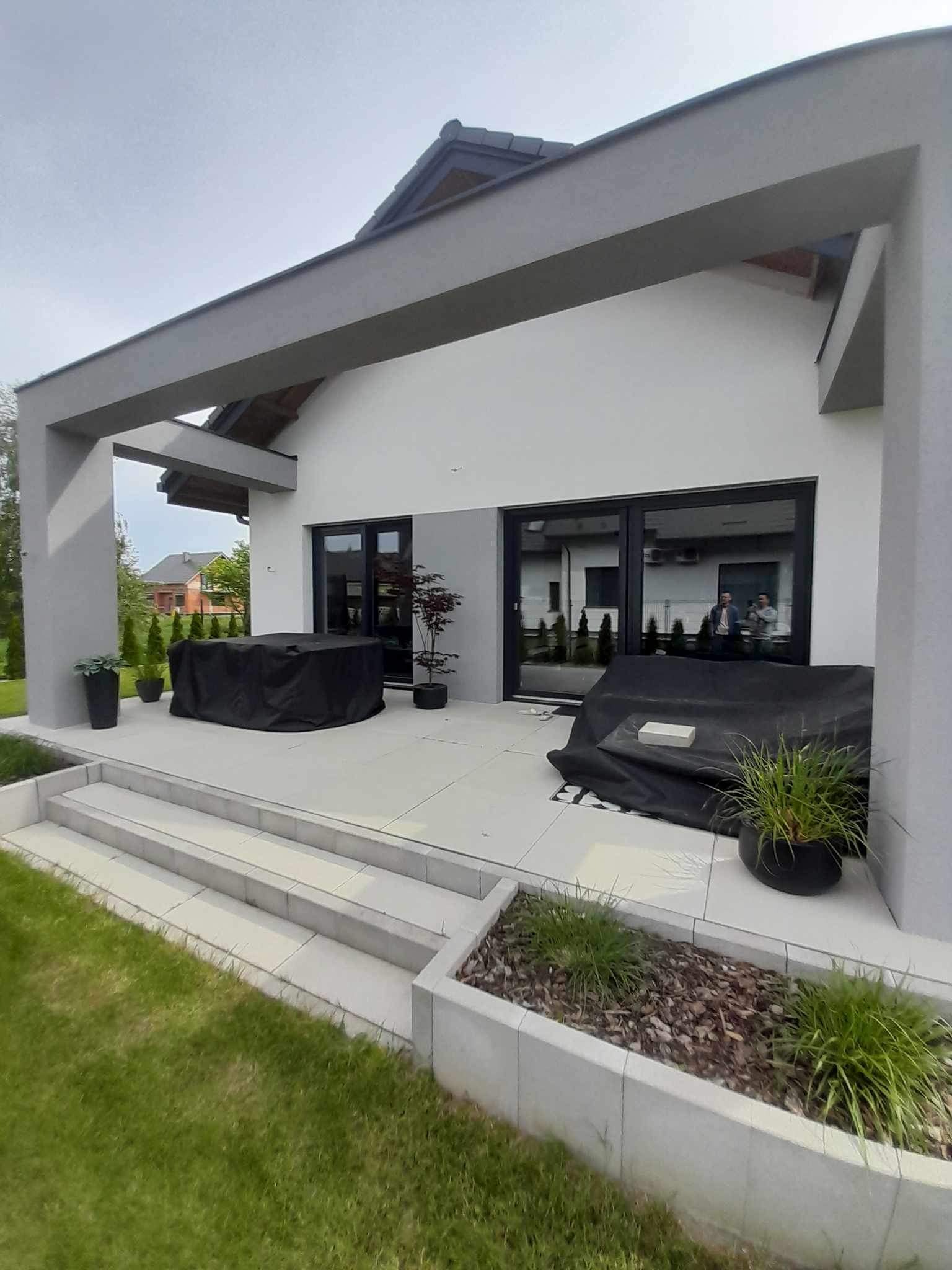 Nowoczesny taras z szarymi płytami, schodami i elementami elewacji, meble ogrodowe przykryte pokrowcem, widoczne donice z roślinami i trawnik