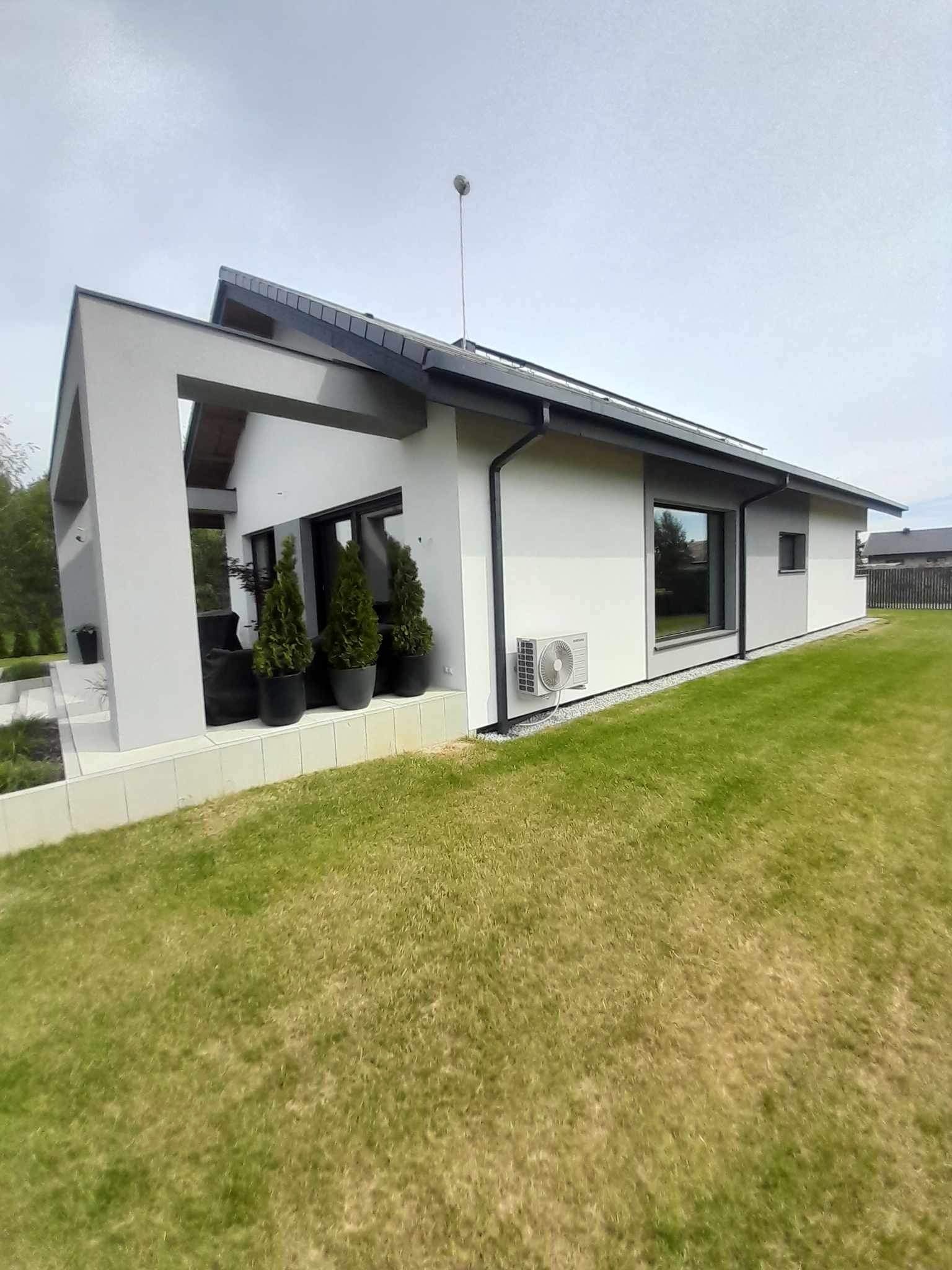 Nowoczesny dom jednorodzinny z jasną elewacją, czarnymi ramami okiennymi i klimatyzatorem na ścianie, otoczony trawnikiem.