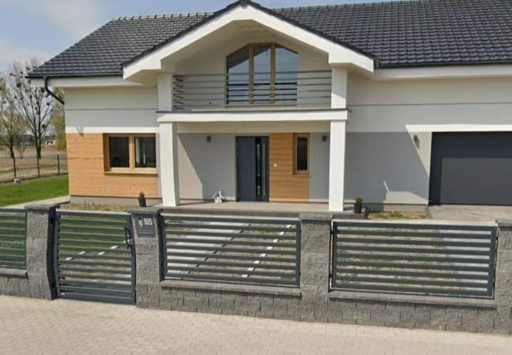 Dwukondygnacyjny dom jednorodzinny z szarą dachówką, białymi ścianami z elementami drewnianej okładziny i ciemnoszarą bramą garażową, ogrodzony nowoczesnym, poziomym ogrodzeniem z szarej kostki...