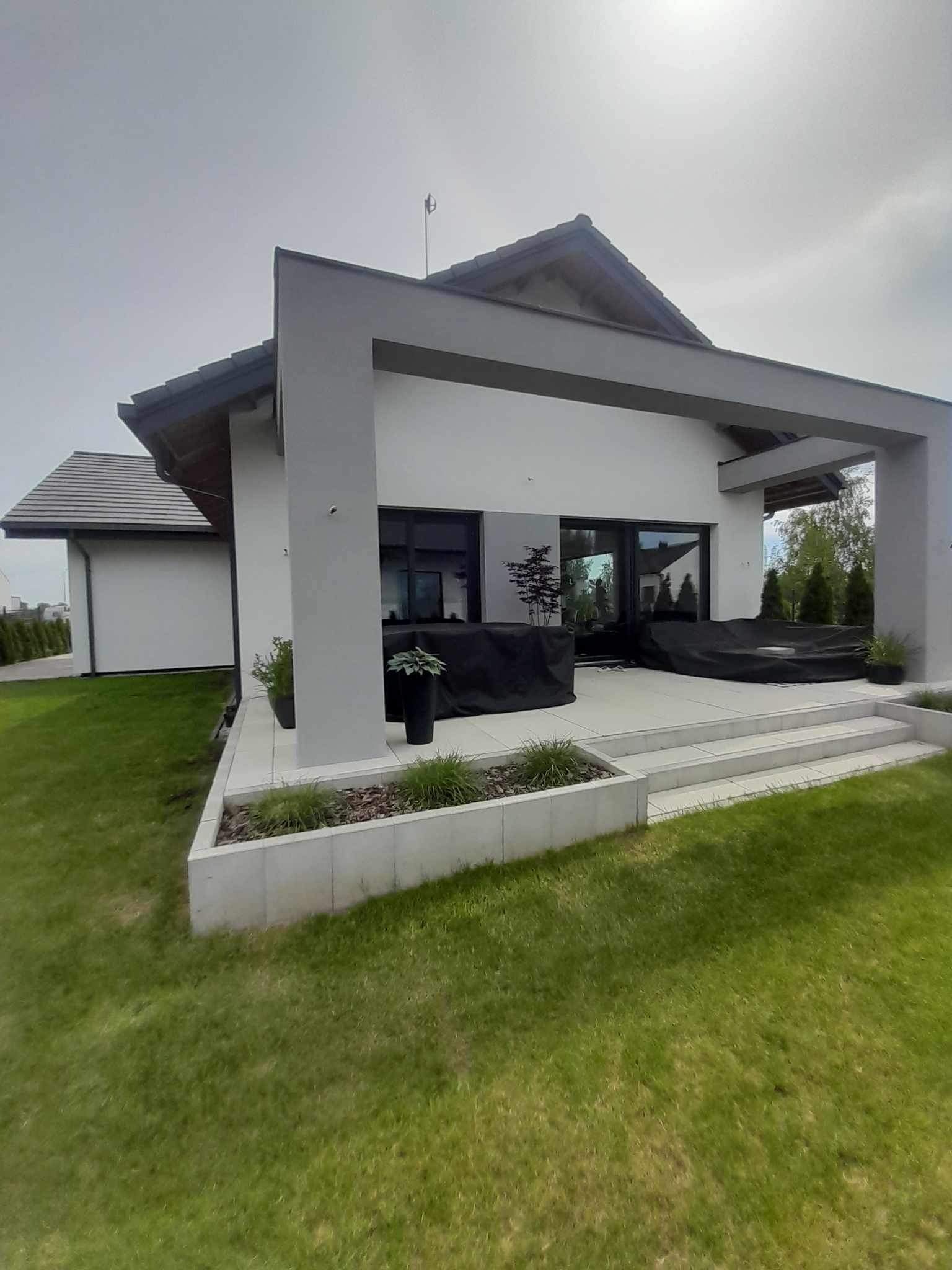 Nowoczesny dom jednorodzinny z białymi ścianami i szarymi elementami architektonicznymi, widok z ogrodu na taras z meblami ogrodowymi przykrytymi czarnymi pokrowcami.