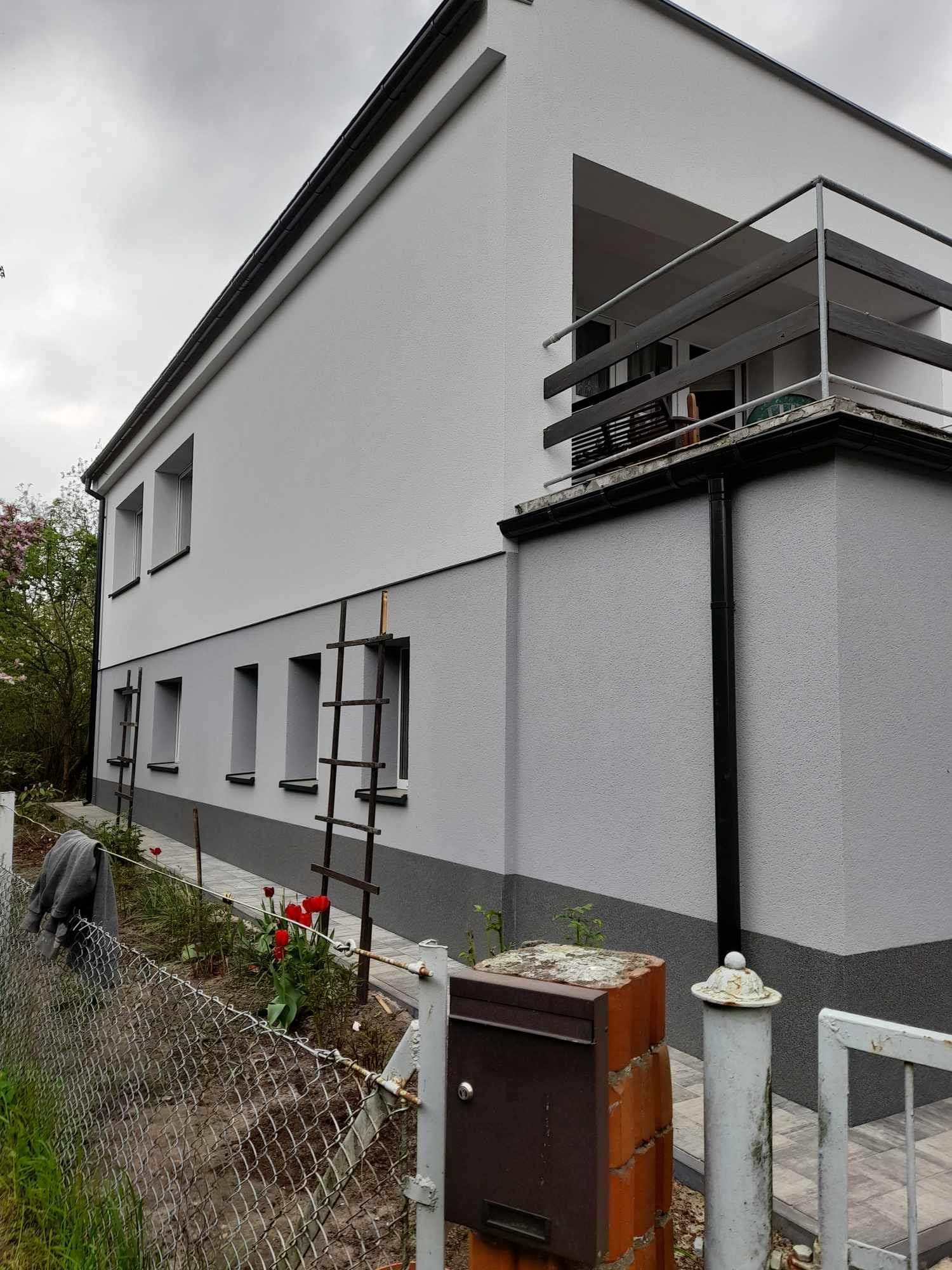 Dwukondygnacyjny dom w odcieniach szarości po remoncie elewacji, widoczne drabiny opierające się o ścianę, skrzynka pocztowa z cegły i metalowe ogrodzenie.