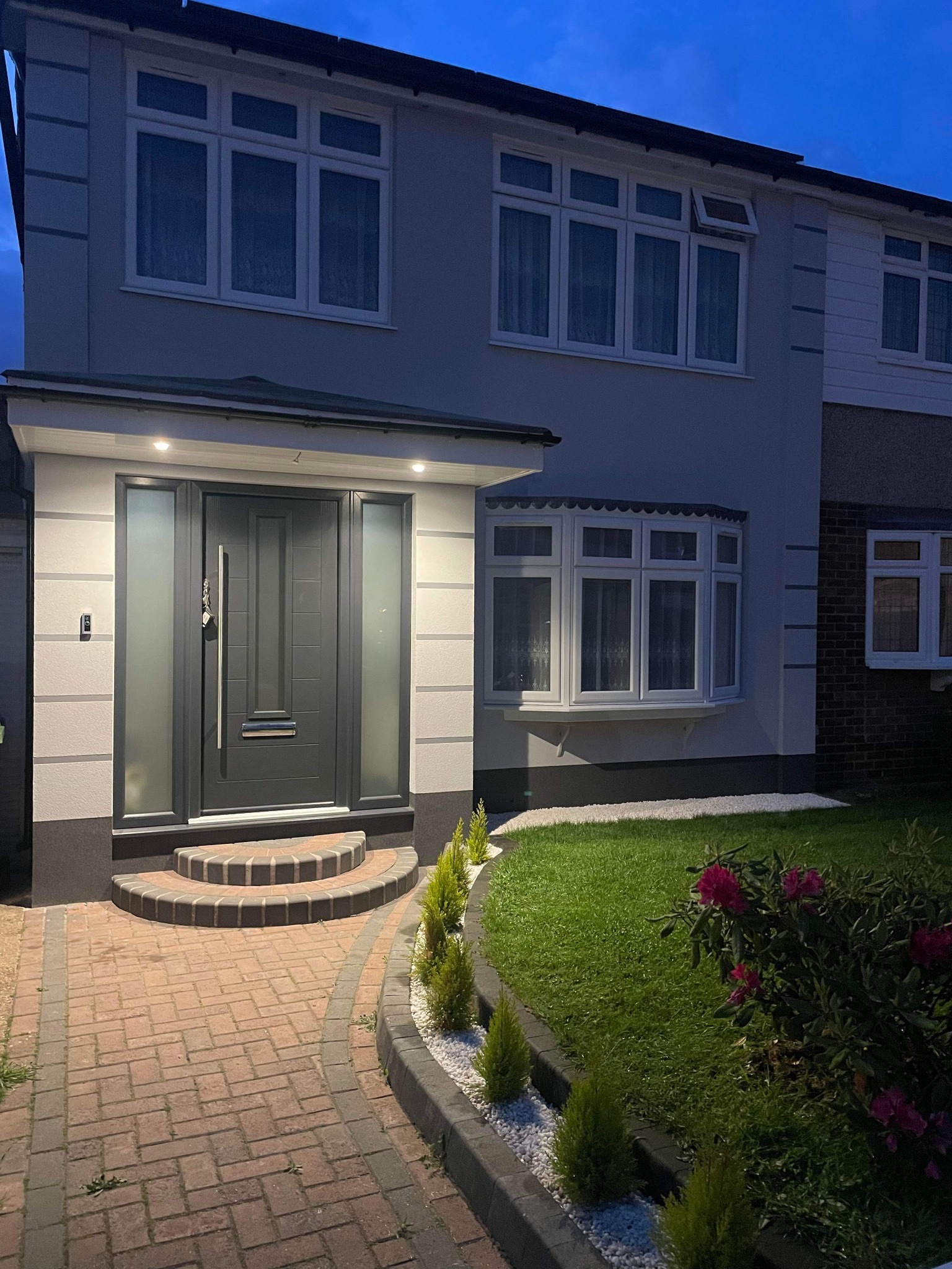 Nowoczesny dom jednorodzinny z szarą elewacją, oświetlonym wejściem z grafitowymi drzwiami i chodnikiem z czerwonej kostki brukowej, widoczne okna z białymi ramami i zadbany trawnik z rabatą kwiatową.