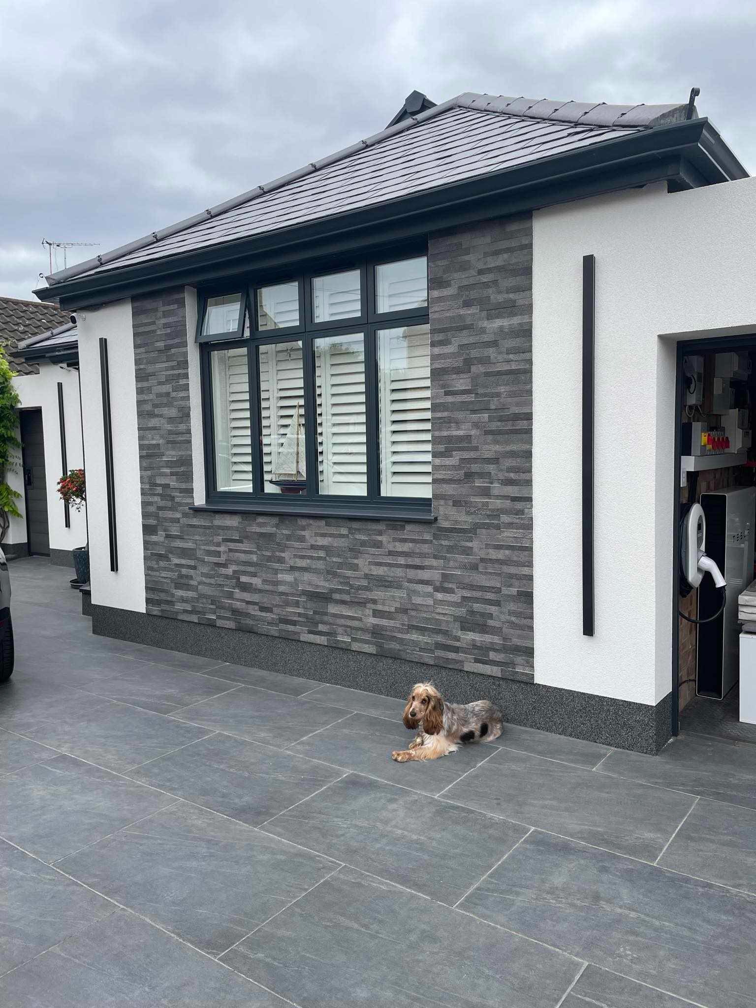 Nowoczesny dom z elewacją z szarej cegły i białym tynkiem, czarnymi detalami oraz leżącym psem rasy Cocker Spaniel na szarym bruku.