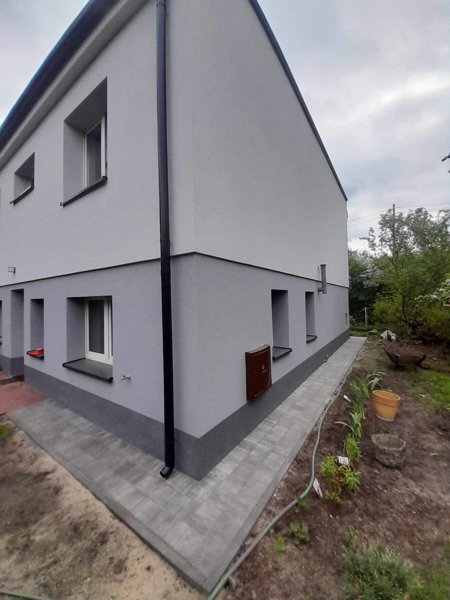 Dwukondygnacyjny dom z szarą elewacją, widoczny fragment ogrodu z kostką brukową i roślinnością, skrzynka na listy na ścianie.