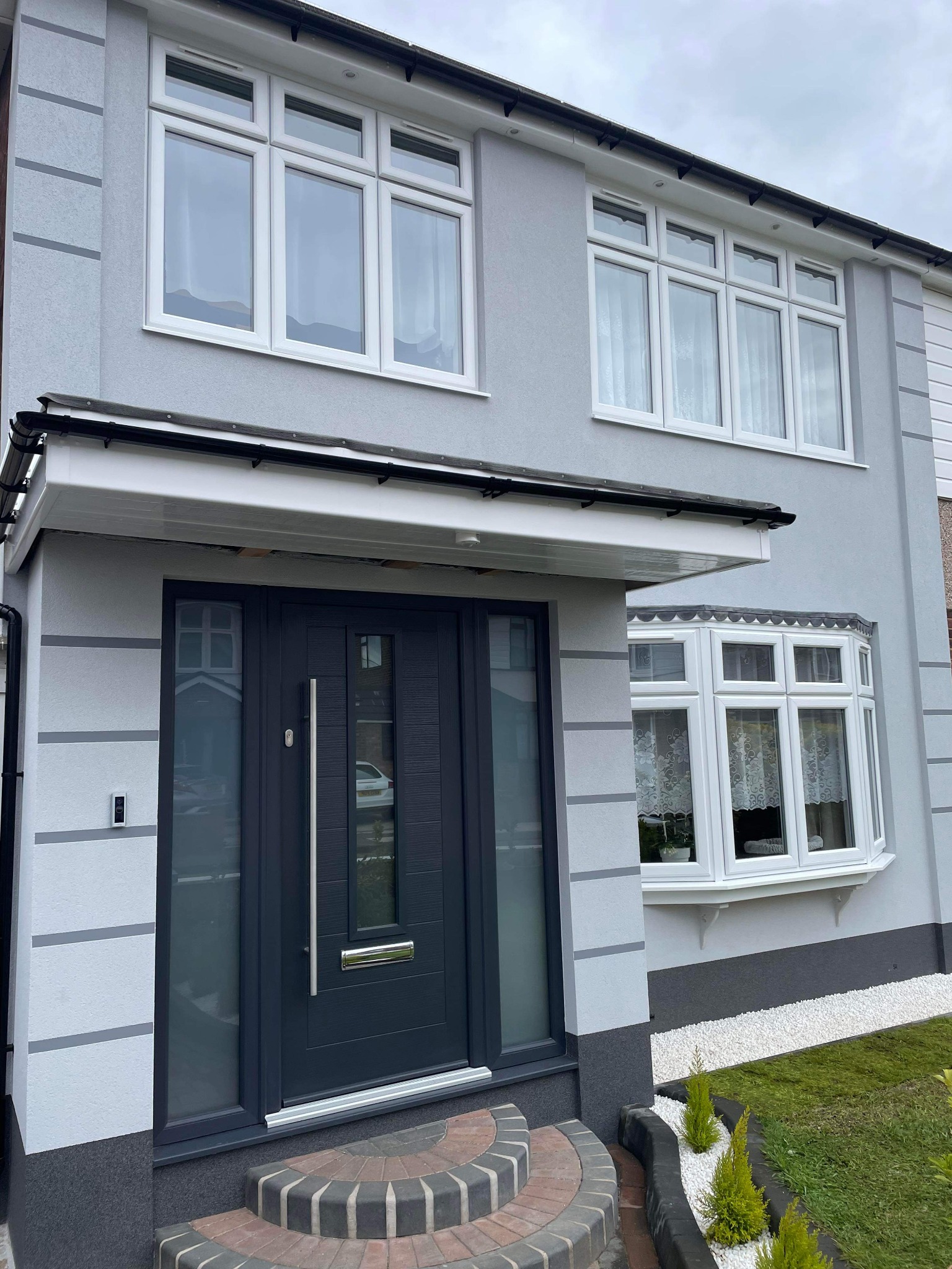 Szary dom jednorodzinny z nową elewacją o strukturze baranka, białą stolarką okienną i ciemnoszarymi drzwiami wejściowymi z daszkiem.