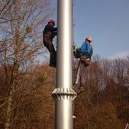 Dwóch monterów w kaskach i uprzężach zabezpieczających pracuje na wysokości przy montażu nowego, metalowego komina na dachu budynku w jesiennej scenerii.