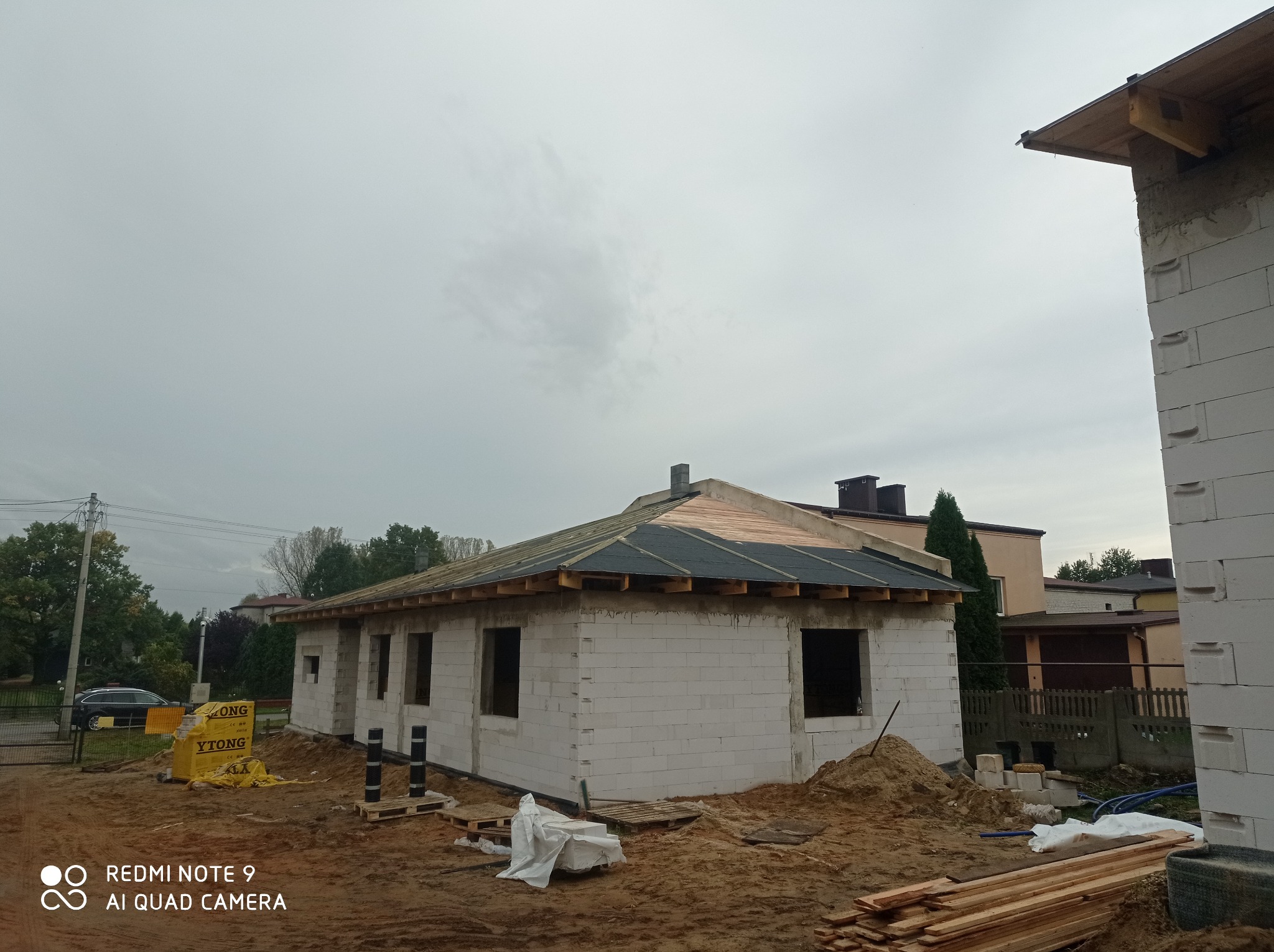 Budowa domu jednorodzinnego w trakcie wznoszenia ścian z bloczków, widoczne pakiety materiałów budowlanych i przygotowane elementy konstrukcji dachu.