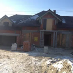 Surowy stan domu w trakcie budowy, z widoczną konstrukcją z czerwonej cegły, częściowo otynkowanymi elementami, ciemną dachówką i stertą piasku na pierwszym planie.