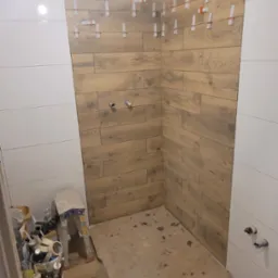 Ściana prysznica w trakcie układania płytek imitujących drewno, z widocznymi krzyżykami dystansowymi, sąsiadująca z fragmentem ściany wyłożonej białymi, poziomymi płytkami. Na podłodze widoczne...