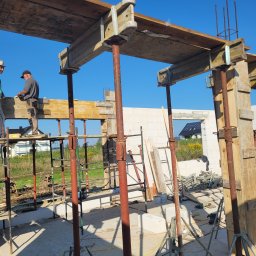 A.R.M. USŁUGI OGÓLNOBUDOWLANE RYSZARD MIKIELEWICZ - Budowa domu jednorodzinnego: szalunek stropu podparty stemplami, widoczni dwaj pracownicy na rusztowaniu, w tle ściany z bloczków i krajobraz z domami.