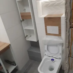 Nowoczesna łazienka w trakcie wykańczania: biała wisząca toaleta z osłoną ochronną, szafka z otwartymi półkami, płytki imitujące drewno oraz dekoracyjna ściana z podświetleniem LED.