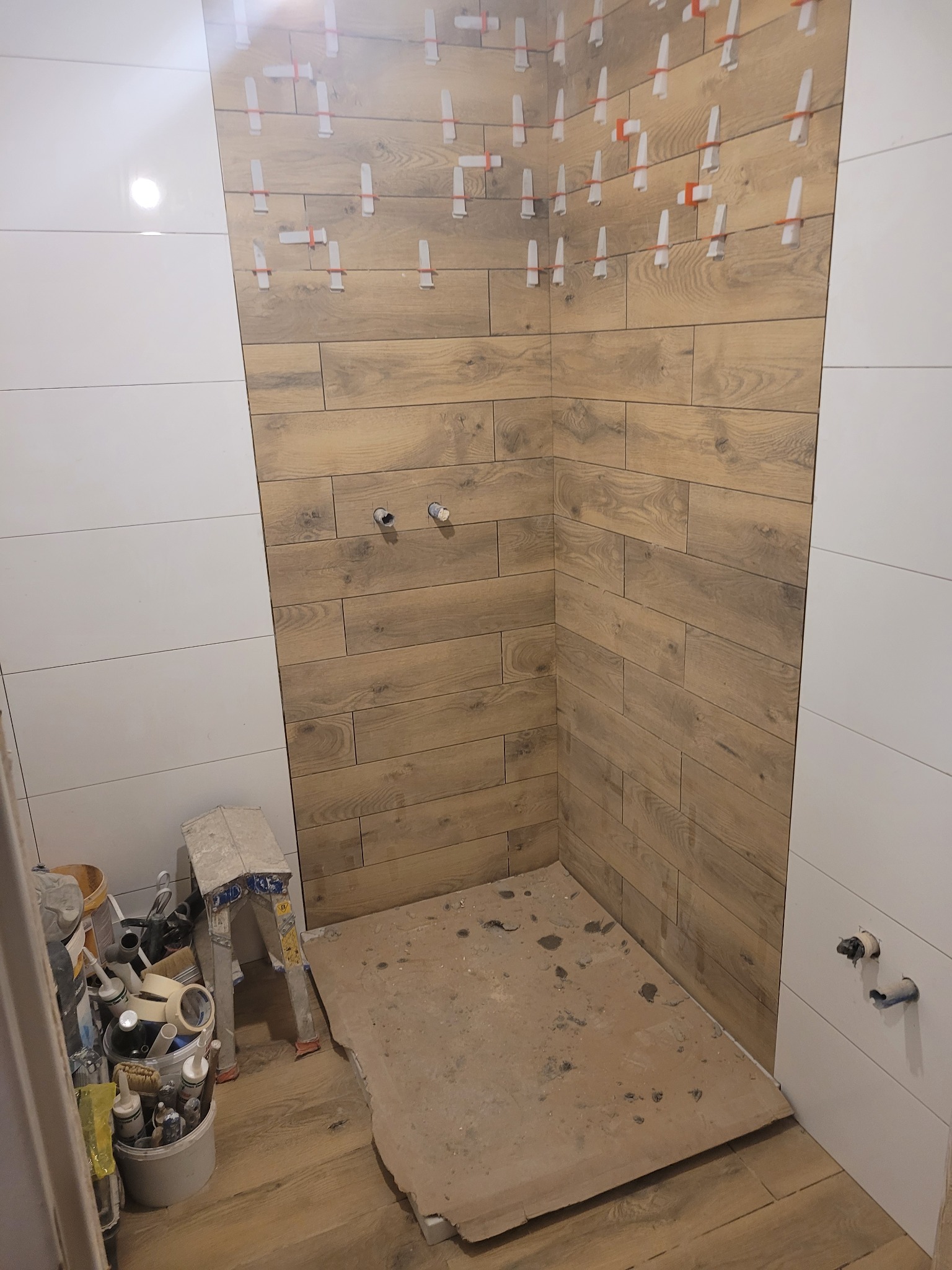 Ściana prysznica w trakcie układania płytek imitujących drewno, z widocznymi krzyżykami dystansowymi, sąsiadująca z fragmentem ściany wyłożonej białymi, poziomymi płytkami. Na podłodze widoczne...