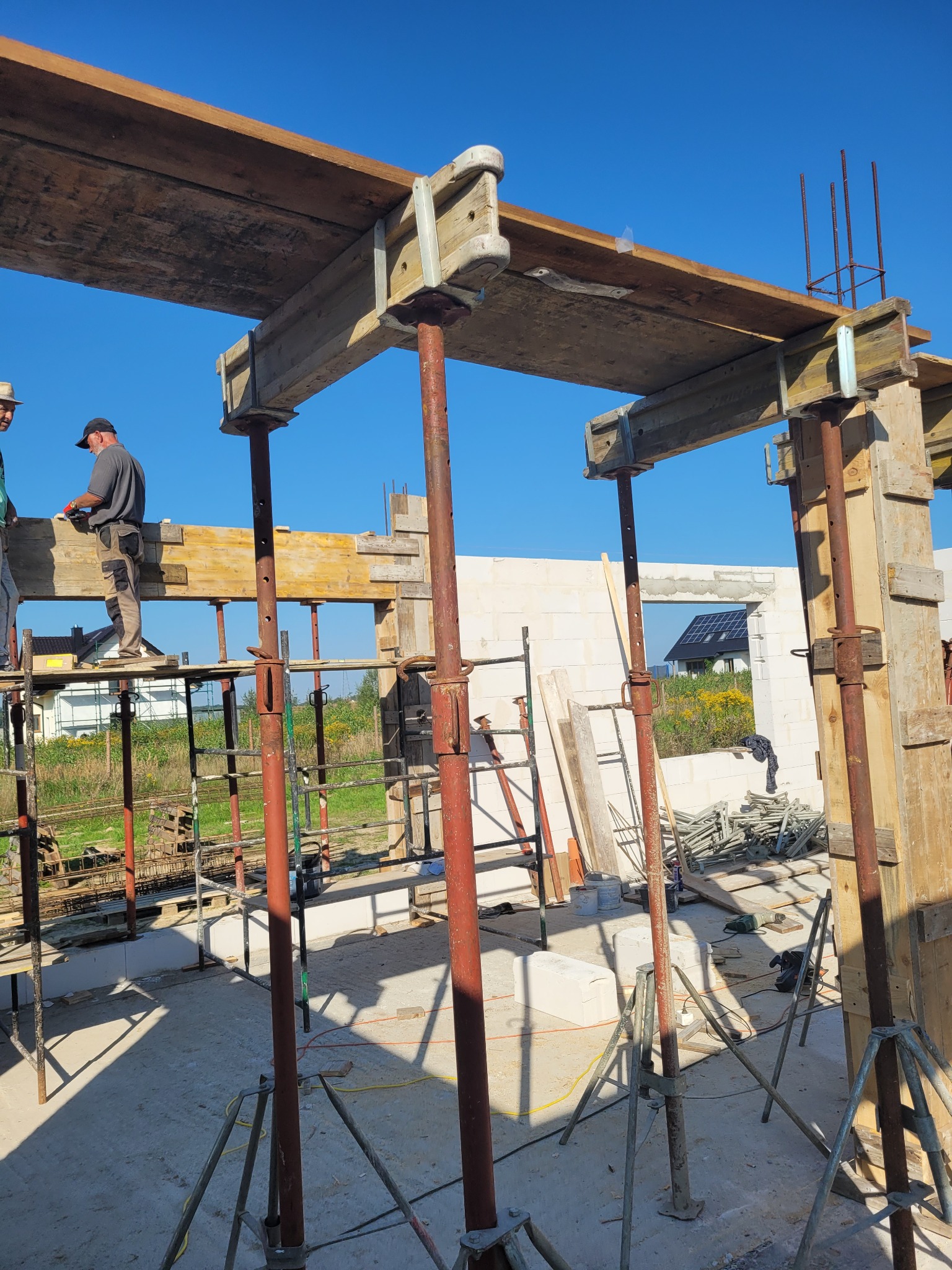 Budowa domu jednorodzinnego: szalunek stropu podparty stemplami, widoczni dwaj pracownicy na rusztowaniu, w tle ściany z bloczków i krajobraz z domami.