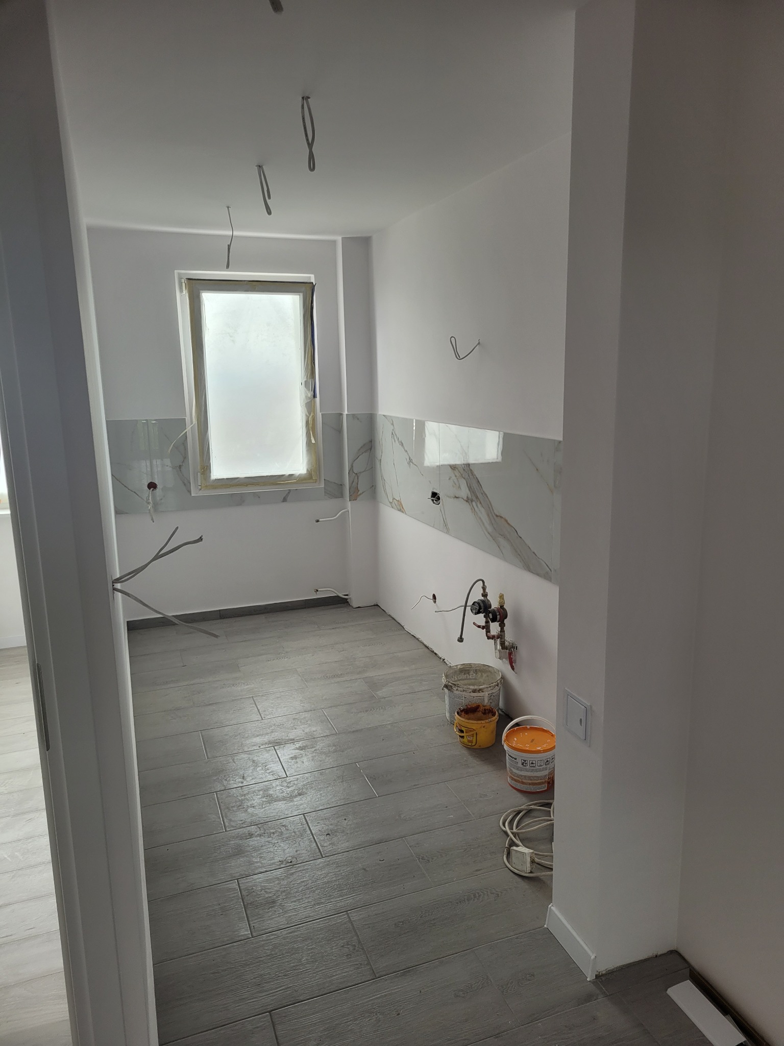Łazienka w trakcie prac wykończeniowych: położona szara podłoga imitująca drewno, fragment ściany z białymi płytkami imitującymi marmur, widoczne puszki z farbą i wystające kable.