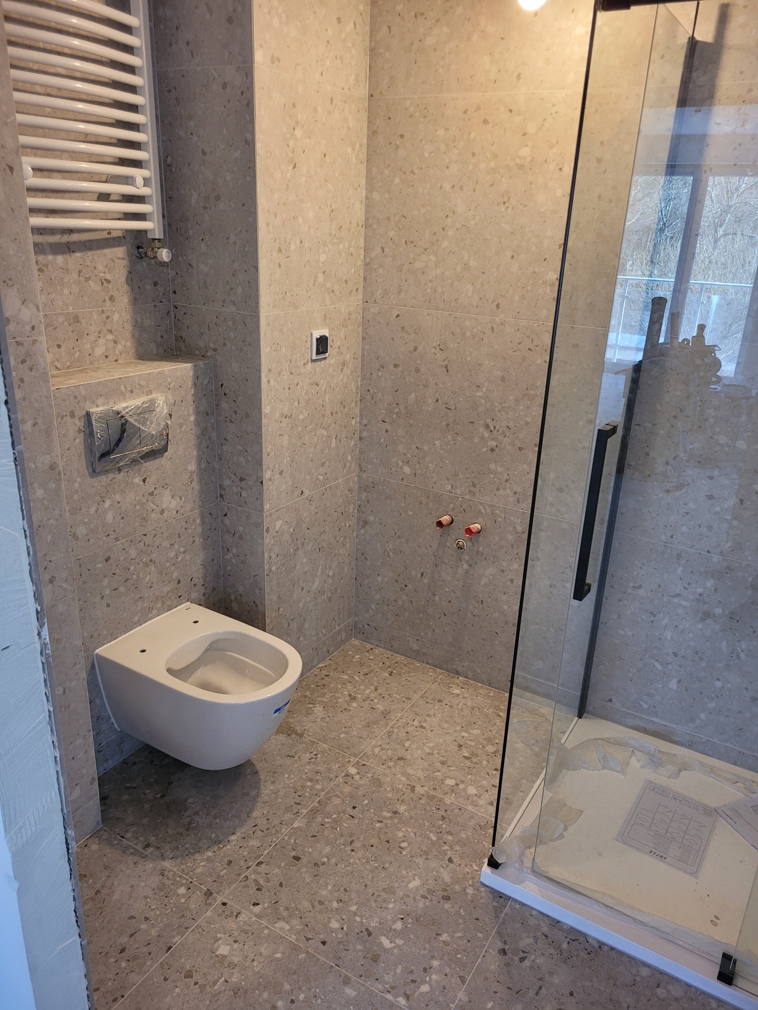 Nowoczesna łazienka w trakcie wykańczania: podwieszana toaleta, grzejnik drabinkowy i kabina prysznicowa ze szklanymi drzwiami, płytki imitujące lastryko na ścianach i podłodze, widoczne przyłącza...