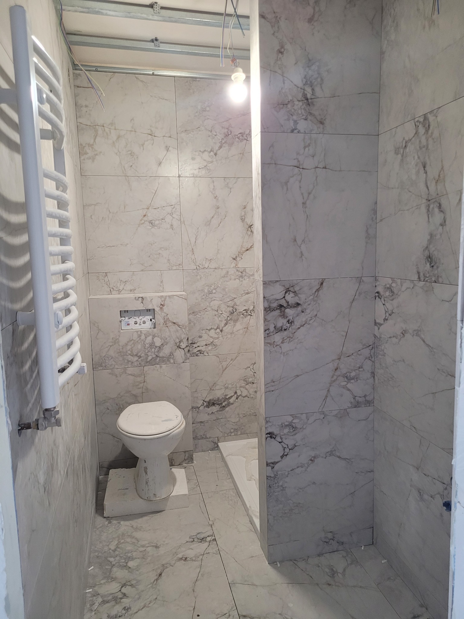 Łazienka w trakcie wykańczania: marmurowe płytki na ścianach i podłodze, wisząca toaleta, grzejnik drabinkowy, sufit podwieszany z wystającymi kablami.
