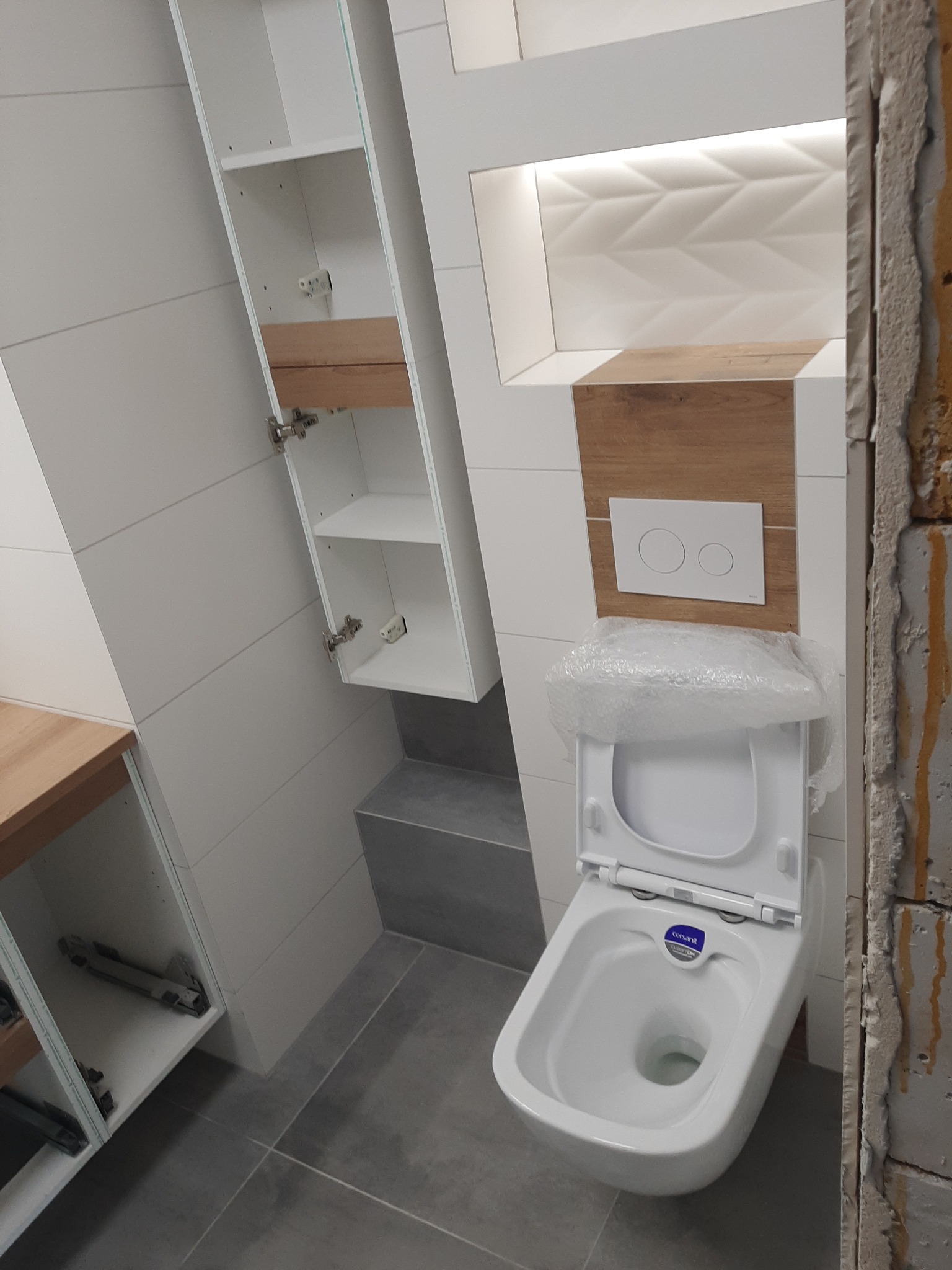 Nowoczesna łazienka w trakcie wykańczania: biała wisząca toaleta z osłoną ochronną, szafka z otwartymi półkami, płytki imitujące drewno oraz dekoracyjna ściana z podświetleniem LED.