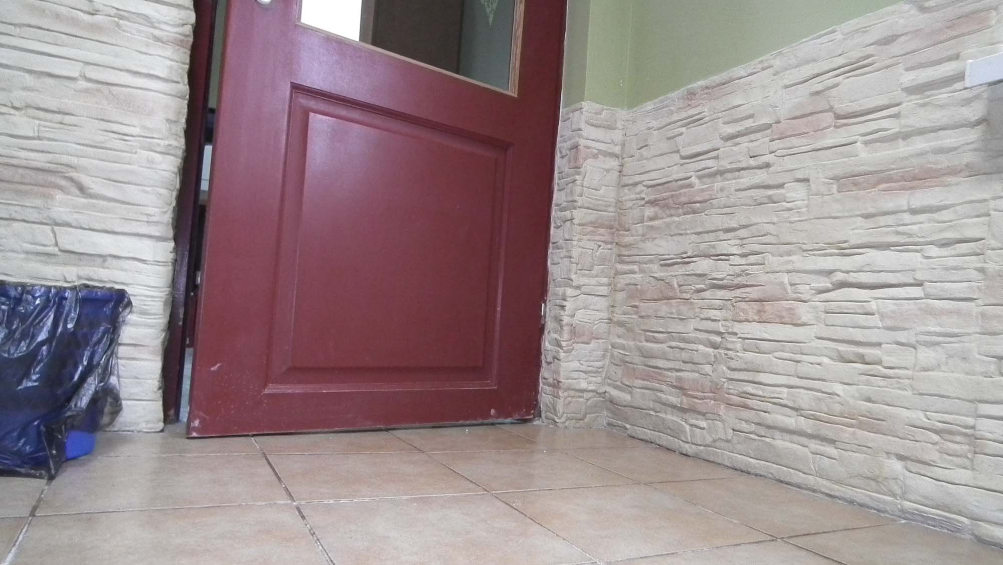 Otwarta, bordowa, drewniana rama drzwiowa z widocznym fragmentem wnętrza, przylegająca do ściany wykończonej panelami imitującymi piaskowiec. Na podłodze beżowe płytki. W lewym dolnym rogu widoczny...