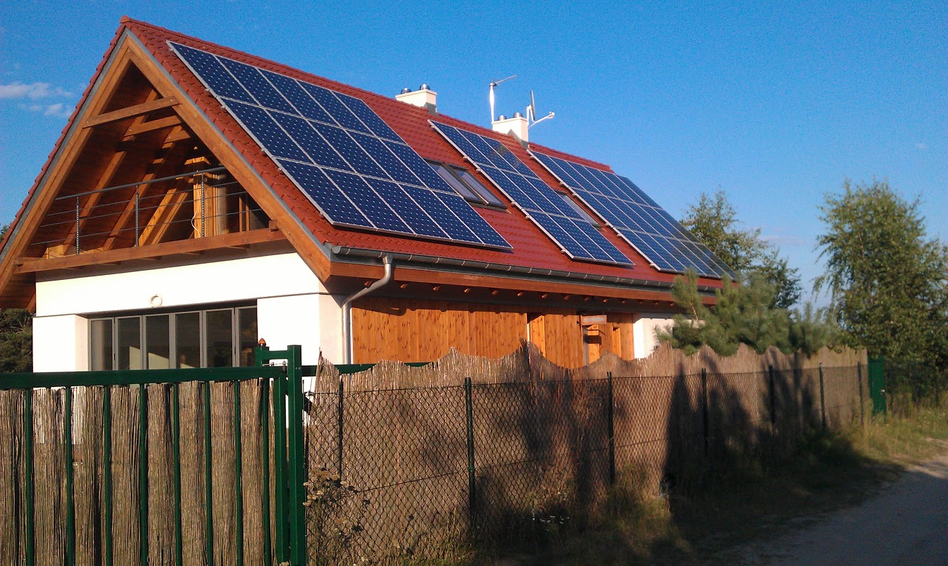 Dom jednorodzinny z panelami fotowoltaicznymi na czerwonym dachu, widok zza ogrodzenia z siatki i trzciny.