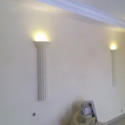 Wnętrze pokoju z dwoma dekoracyjnymi kolumnami oświetleniowymi na ścianie, widoczny fragment sufitu z listwą dekoracyjną i zabezpieczone folią malarską krzesło.