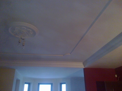 Biały sufit z dekoracyjnym rozetą i listwami, widoczne niedokończone prace malarskie, ściana pomalowana na czerwono w rogu pomieszczenia.