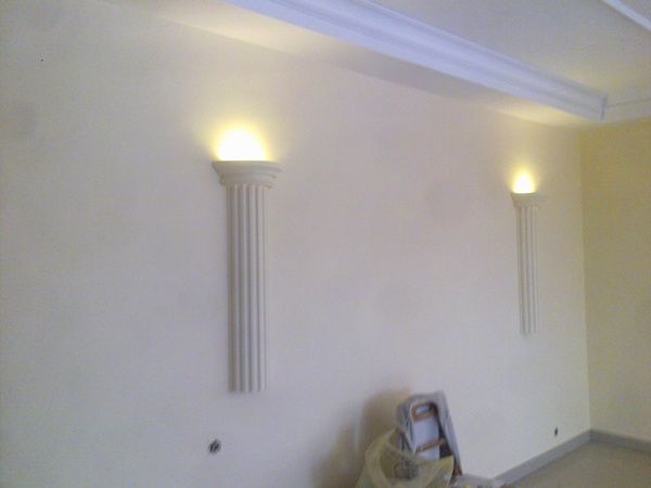 Wnętrze pokoju z dwoma dekoracyjnymi kolumnami oświetleniowymi na ścianie, widoczny fragment sufitu z listwą dekoracyjną i zabezpieczone folią malarską krzesło.