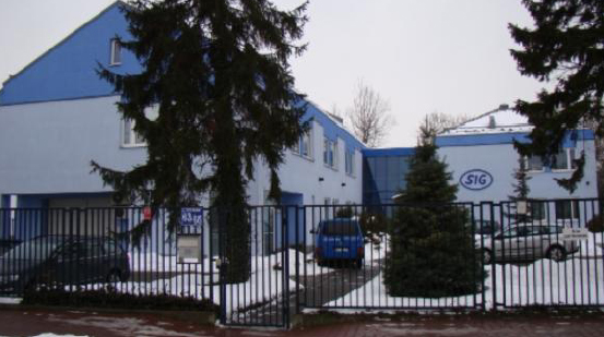 Dwupiętrowy budynek biurowy z niebieskimi akcentami, widoczny zza czarnego metalowego ogrodzenia w zimowej scenerii z resztkami śniegu, z logo 'SIG' na elewacji.