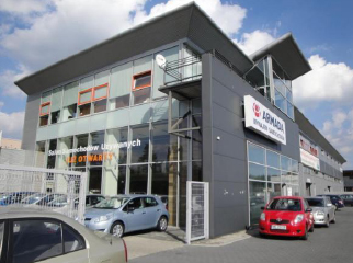 Nowoczesny, dwukondygnacyjny budynek biurowy z szarą elewacją, dużymi oknami i logo firmy Armada na fasadzie. Przed budynkiem zaparkowane samochody.