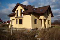 Żółty dom jednorodzinny z brązowym dachem i oknami, sfotografowany w pochmurny dzień z perspektywy lekko z dołu, z widoczną suchą trawą na pierwszym planie.