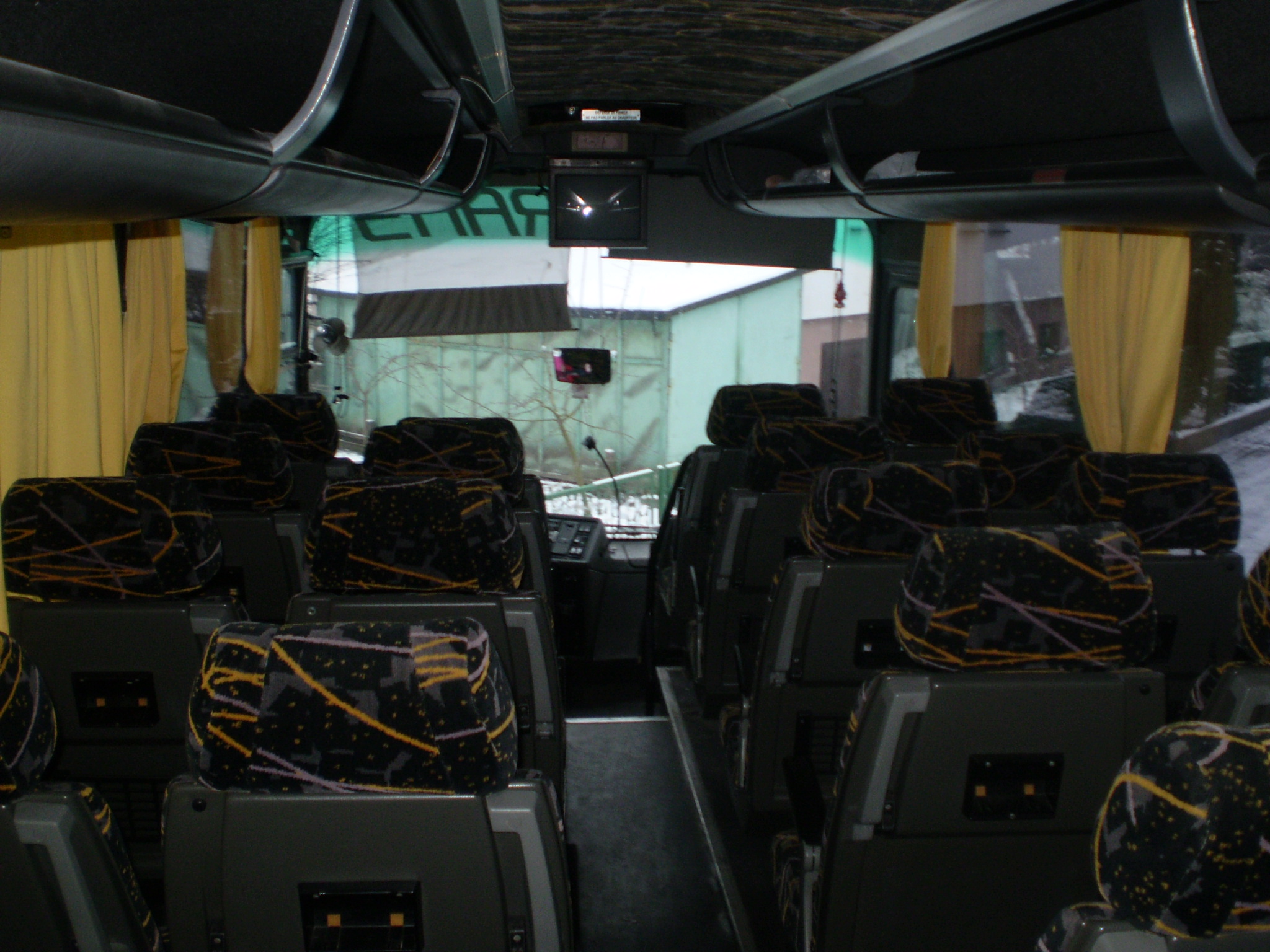 Wnętrze autokaru z rzędami foteli o wzorzystej tapicerce, żółte zasłonki przy oknach, widoczny kokpit kierowcy i monitory pod sufitem.