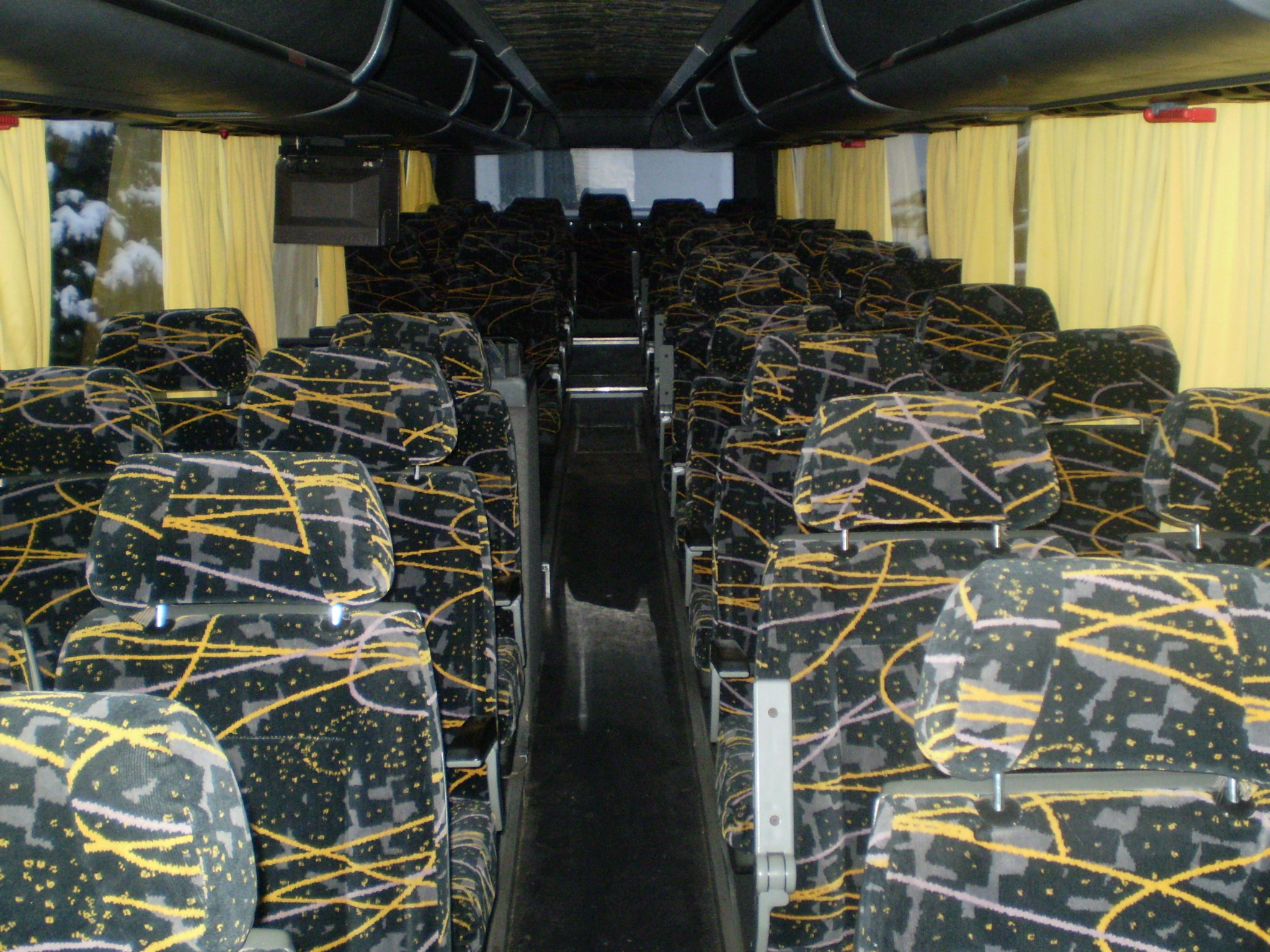Wnętrze autokaru z rzędami foteli obitych czarną tapicerką w geometryczne wzory, widok z przodu wzdłuż korytarza, żółte zasłony w oknach.