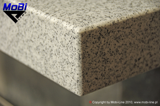 Zbliżenie na narożnik blatu mineralno-akrylowego solid surface o jasnej, nakrapianej fakturze, z widocznym logo producenta w lewym górnym rogu.