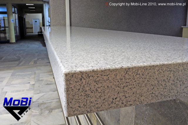 Jasny blat mineralno-akrylowy o drobnej fakturze, z widocznym zaokrąglonym brzegiem, zamontowany w przestrzeni publicznej w Makowie Podhalańskim. W tle widoczny korytarz z marmurową posadzką.