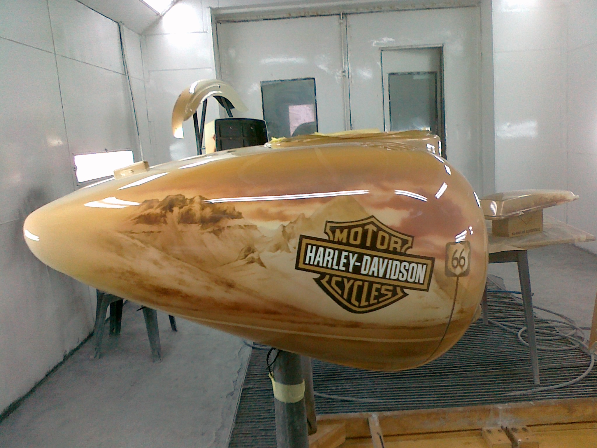 Lakierowany zbiornik paliwa motocykla Harley-Davidson z motywem górskim i Route 66, w kabinie lakierniczej.