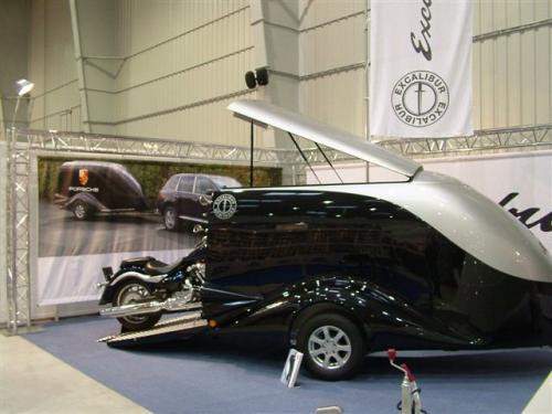 Czarny, niestandardowy pojazd z srebrnym dachem, przypominający połączenie motocykla i przyczepy, na tle baneru reklamowego z przyczepą Porsche i SUV-em, prezentowany na targach.