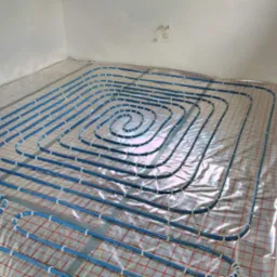 Niebieskie rury ogrzewania podłogowego ułożone spiralnie na folii z siatką w pomieszczeniu w Polance, widoczne białe ściany i wystające rurki.